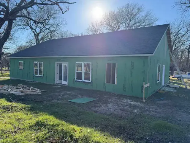 New house under construction with green siding, windows, and black roof, set in a grassy outdoor area on a sunny day.