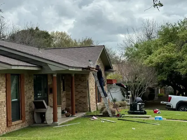 Person on a ladder working on a roof gutter of a house; tools and materials on the ground.