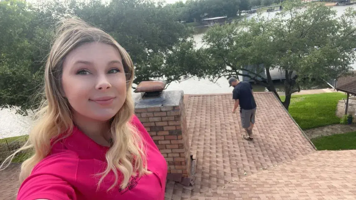 Woman in pink shirt takes a selfie on a brick roof; man walks on the roof, water and trees in the background.