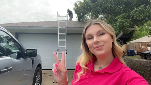 Woman making a peace sign in front of a house, someone on a ladder cleaning the roof.