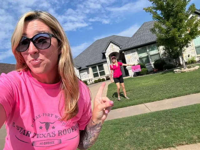 Woman in pink shirt and sunglasses points to another person in front of a house.