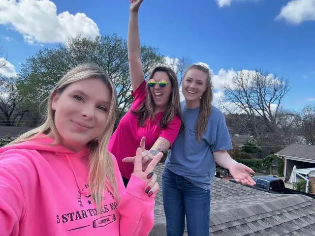 Three people on a rooftop, smiling. One in pink hoodie, one in pink shirt, one in blue shirt. Sunny day, trees in background.