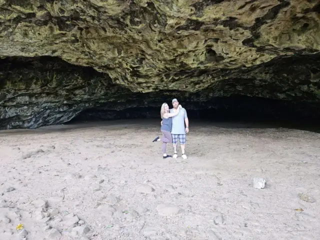 Two people embrace in a large, dark cave with a sandy floor.