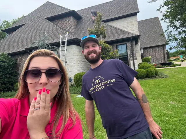 Woman with sunglasses blows a kiss, man smiles. Roofers on a house with a ladder, green lawn.