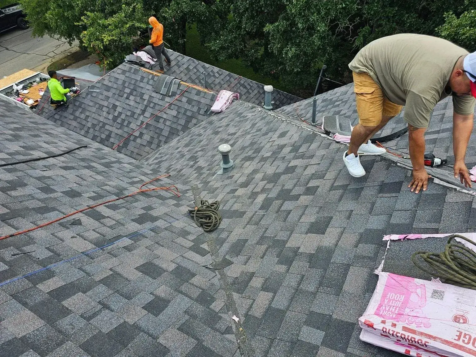 Roofers working on a gray shingle roof. One kneels, others stand, with tools and materials visible.