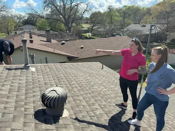 Three people on a brown shingle roof, one points, others work. Sunny day.