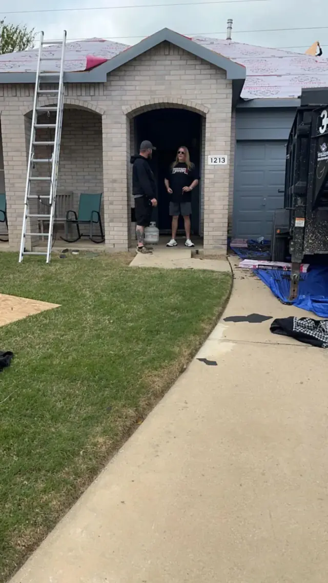 Two people standing in a home's entryway as roof work is done; a ladder and a truck are visible.