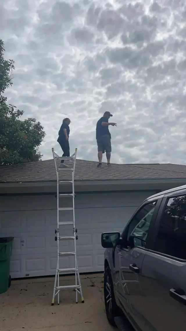 Two people on a roof near a ladder and a car under a cloudy sky.